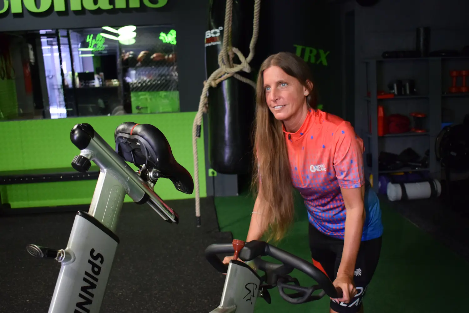 Spinning edzés résztvevői tekernek a TFWG12 sportstúdióban, lendületes zenére, edzői irányítással, állva és ülve váltakozó gyakorlatokkal.